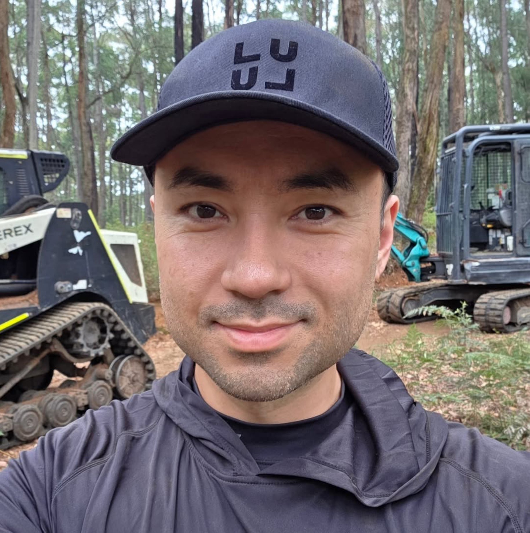Alexander Wong in a cap, smiling, in the bush with farm machinery behind him